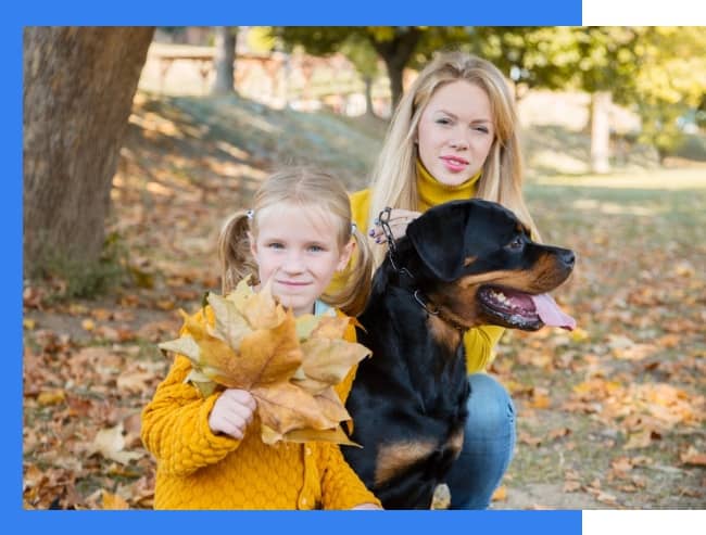 A woman and a young girl with blonde hair pose outdoors with a large black dog; the girl holds yellow autumn leaves.