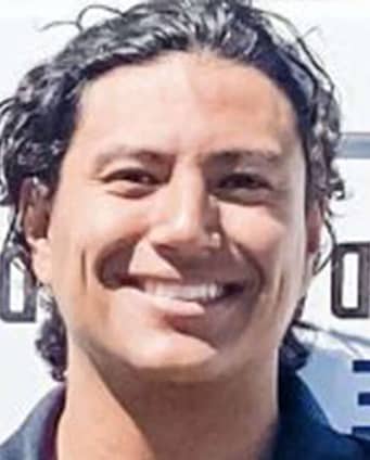 A person with wavy dark hair is smiling at the camera in front of a blurred background with partial text.