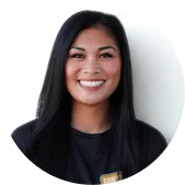 A woman with long black hair and a black shirt stands smiling in front of a plain, light-colored background.