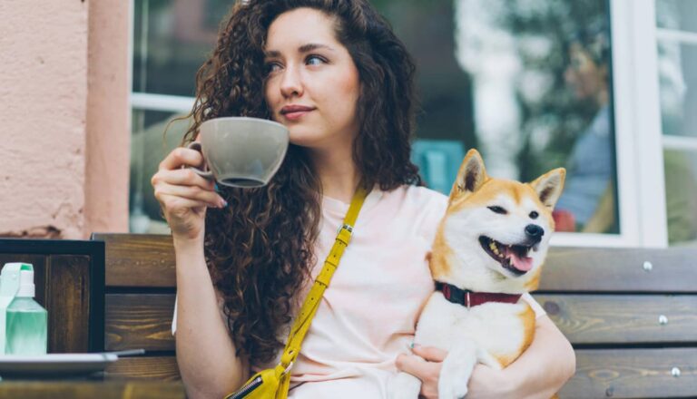 A woman with curly hair sits on a bench, holding a cup and a smiling Shiba Inu dog in her lap.