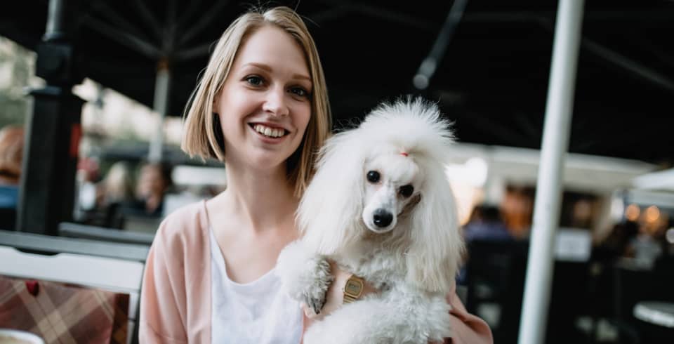 A woman with blonde hair smiles while holding a small white poodle in her arms at an outdoor café.