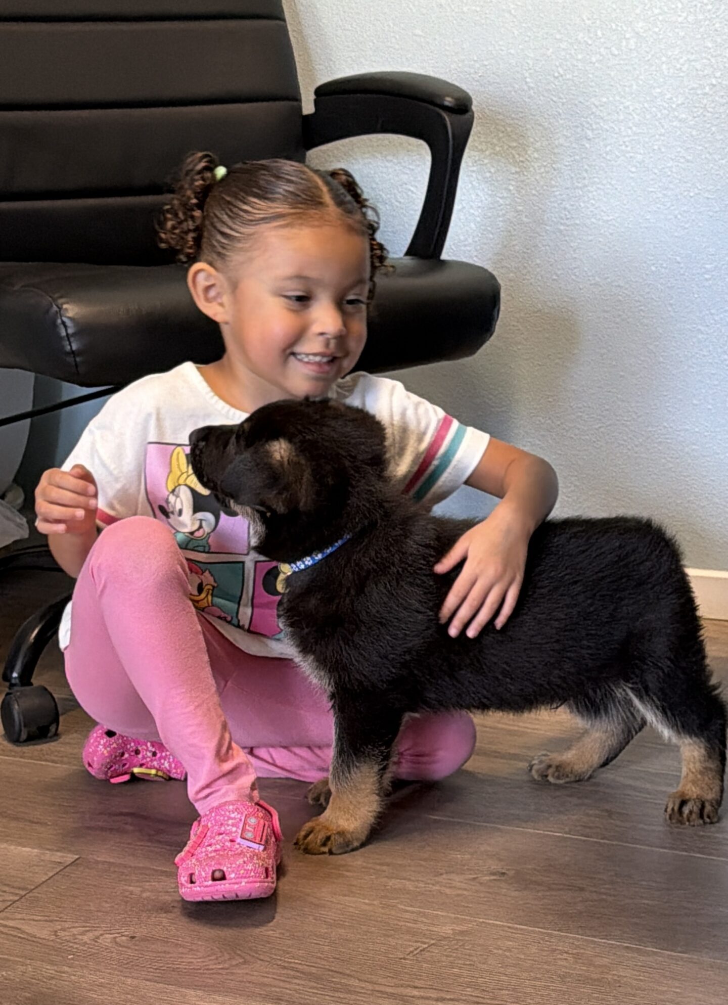A young girl sitting on the floor pets a black and tan puppy indoors, with an office chair in the background.
